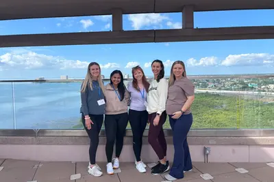 Five people stand together on an outdoor terrace, smiling at the camera with a waterfront and city skyline in the background.