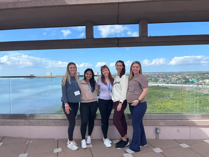 Five people stand together on an outdoor terrace, smiling at the camera with a waterfront and city skyline in the background.