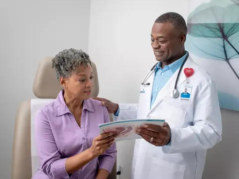 Doctor in white coat and stethoscope explaining a medical diagram to a seated patient.