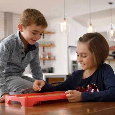 Two children playing with a red toy on a wooden table in a kitchen.