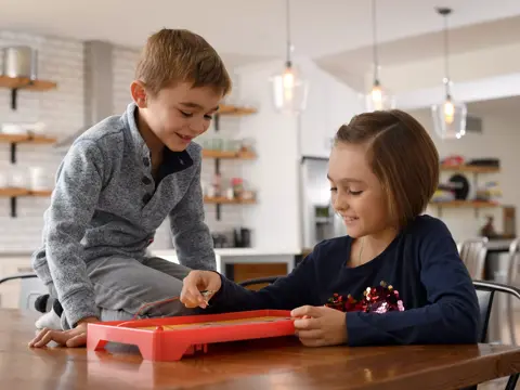 Two children playing with a red toy on a wooden table in a kitchen.