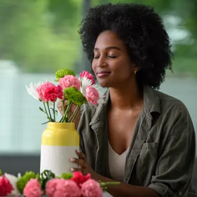A woman with afro hair holds a vase of pink and green flowers, smiling.