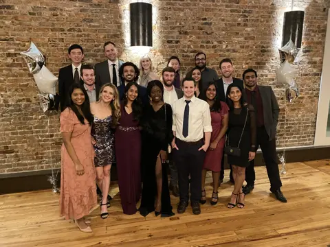 Group dressed in formal attire posing indoors against a brick wall with star-shaped balloons.
