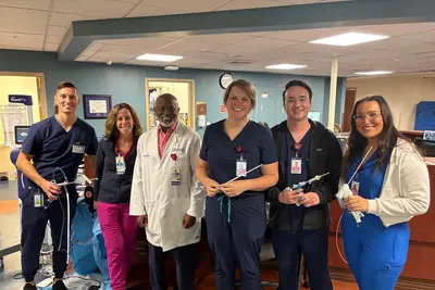 Six healthcare professionals in scrubs and a white coat pose together in a clinical work area, several holding medical instruments and smiling at the camera