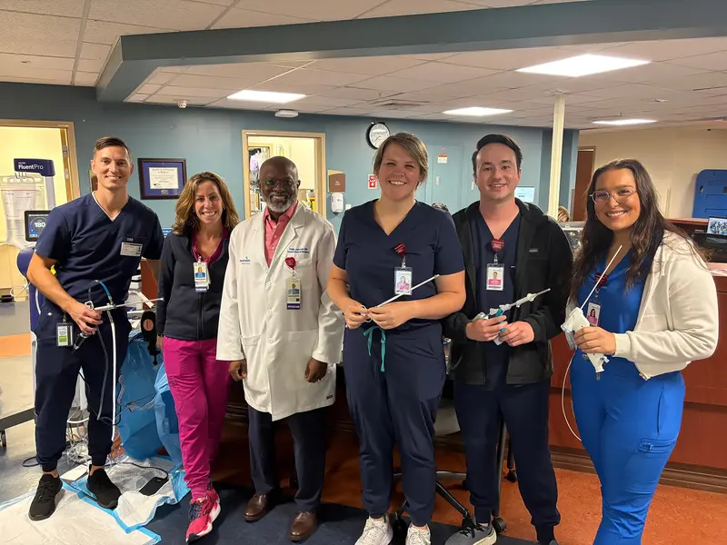 Six healthcare professionals in scrubs and a white coat pose together in a clinical work area, several holding medical instruments and smiling at the camera