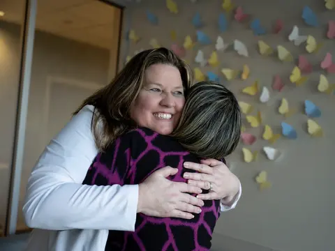 Two women embrace in a warm hug, smiling brightly against a backdrop of colorful butterfly decorations.
