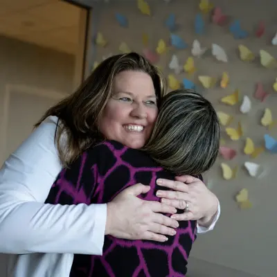 Two women embrace in a warm hug, smiling brightly against a backdrop of colorful butterfly decorations.