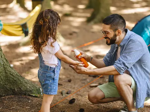 Father kneeling outdoors applying bug spray to his young daughter's hand during a camping trip