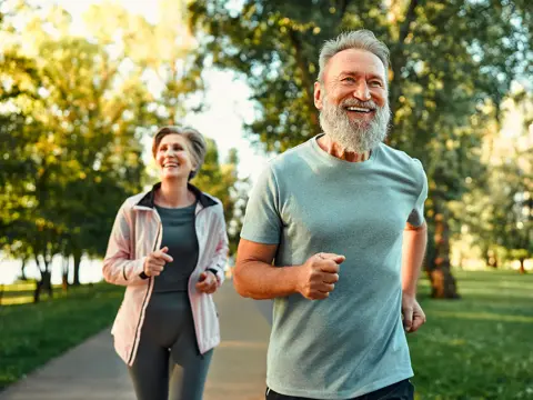 Smiling older couple jogging on a sunlit park path, trees and greenery blurred in the background.