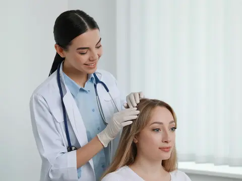 A healthcare provider in a white coat and stethoscope examining a patient's scalp during a hair treatment consultation in a clinical setting