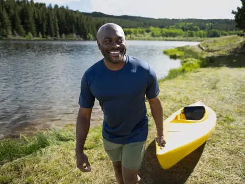 A smiling man stands by a lake with a yellow kayak, surrounded by greenery and trees.