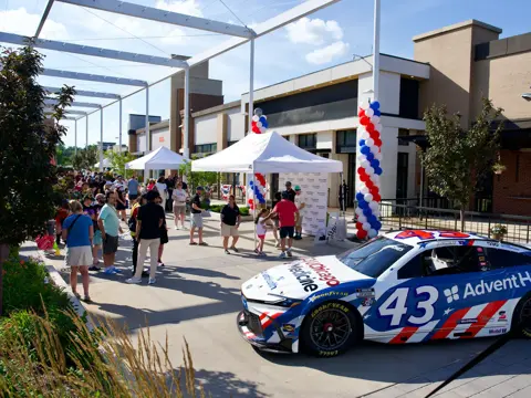 AdventHealth-branded NASCAR race car displayed at an outdoor community event with white tents, red, white, and blue balloon columns, and people gathering nearby.