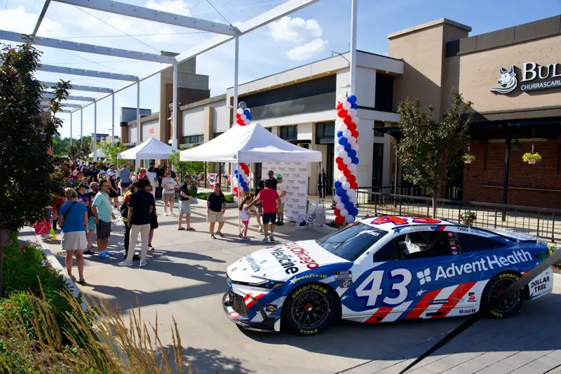 AdventHealth-branded NASCAR race car displayed at an outdoor community event with white tents, red, white, and blue balloon columns, and people gathering nearby.