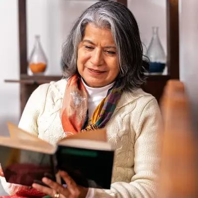 Gray-haired woman reads a book indoors; cream sweater, colorful scarf; shelves and vases behind