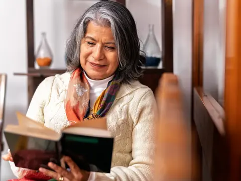 Gray-haired woman reads a book indoors; cream sweater, colorful scarf; shelves and vases behind
