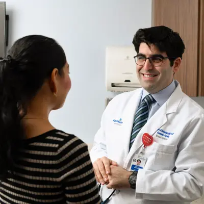 Medical professional in white coat with AdventHealth branding smiling while consulting with a patient in a clinical exam room