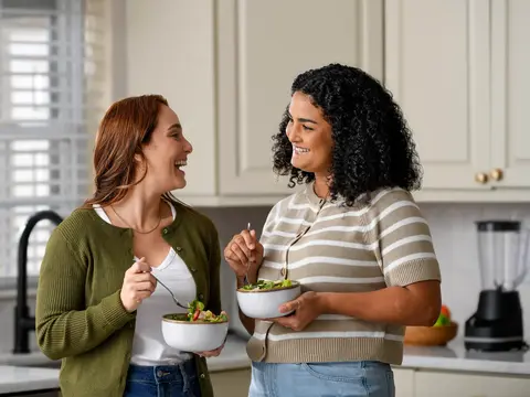Two women smiling and eating salads together in a bright modern kitchen