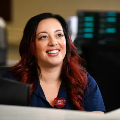 Smiling nurse in navy blue scrubs with red-highlighted hair engaging with a colleague at a nurses station in a hospital setting