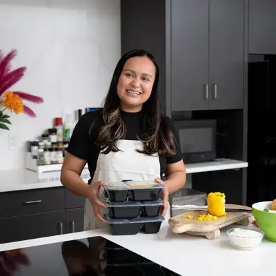 A woman in a kitchen holding meal prep containers, surrounded by ingredients and kitchenware.