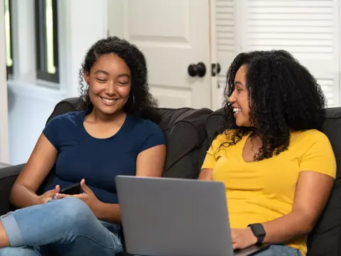 Two women sitting on a couch, smiling and using a laptop.