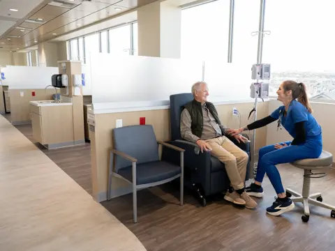 Healthcare professional in blue scrubs seated on a stool engaging with an elderly male patient in an infusion chair in a bright modern medical facility with large windows