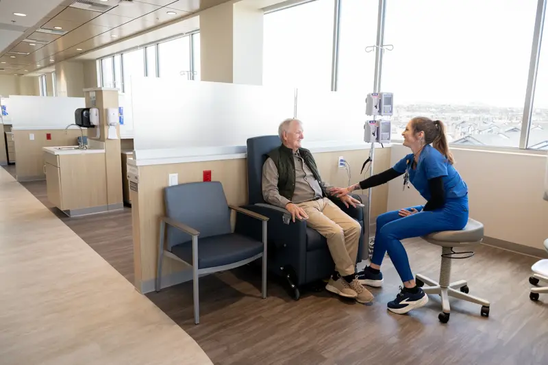 Healthcare professional in blue scrubs seated on a stool engaging with an elderly male patient in an infusion chair in a bright modern medical facility with large windows